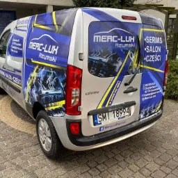 Srebrny samochód dostawczy Mercedes-Benz z niebiesko-żółtą grafiką reklamową firmy Merc-Lux Rzymski, prezentujący usługi serwisowe i sprzedaż części samochodowych, zaparkowany na brukowanym...