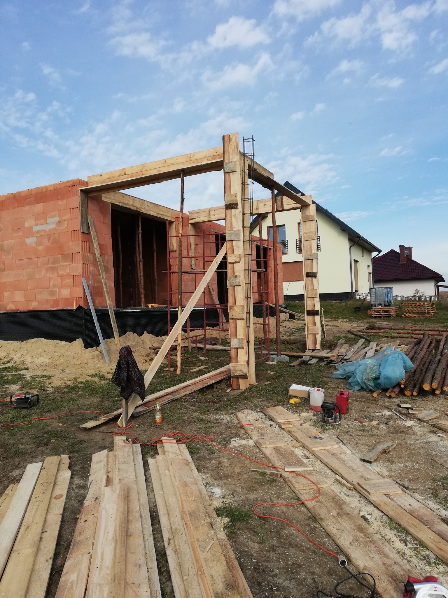 Budowa domu w trakcie: ściana z czerwonej cegły, drewniana konstrukcja szkieletowa, widoczne materiały budowlane i narzędzia na placu budowy, w tle dom jednorodzinny.