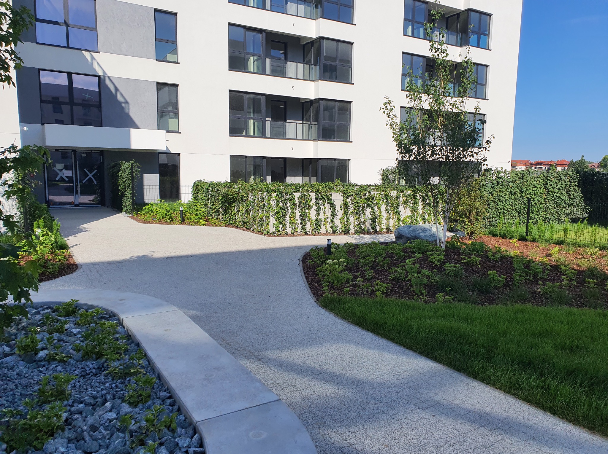 Nowoczesny budynek z zadbanym terenem zielonym, wijącym się chodnikiem z kostki brukowej i starannie dobraną roślinnością pnącą się po ogrodzeniu.