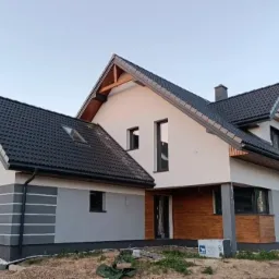 Nowoczesny dom jednorodzinny z szarą elewacją i drewnianymi akcentami, czarny dach z lukarną, widoczne materiały budowlane na posesji.