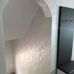 Korytarz ze schodami i łukowatym przejściem, ściany pokryte dekoracyjną szpachlą w odcieniach szarości, obok czarny panel z wieszakami i siedziskiem.