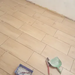 Ułożone płytki podłogowe imitujące drewno, z krzyżykami dystansowymi, widoczny fragment ściany w trakcie prac wykończeniowych, kuweta z krzyżykami i narzędzia do fugowania.