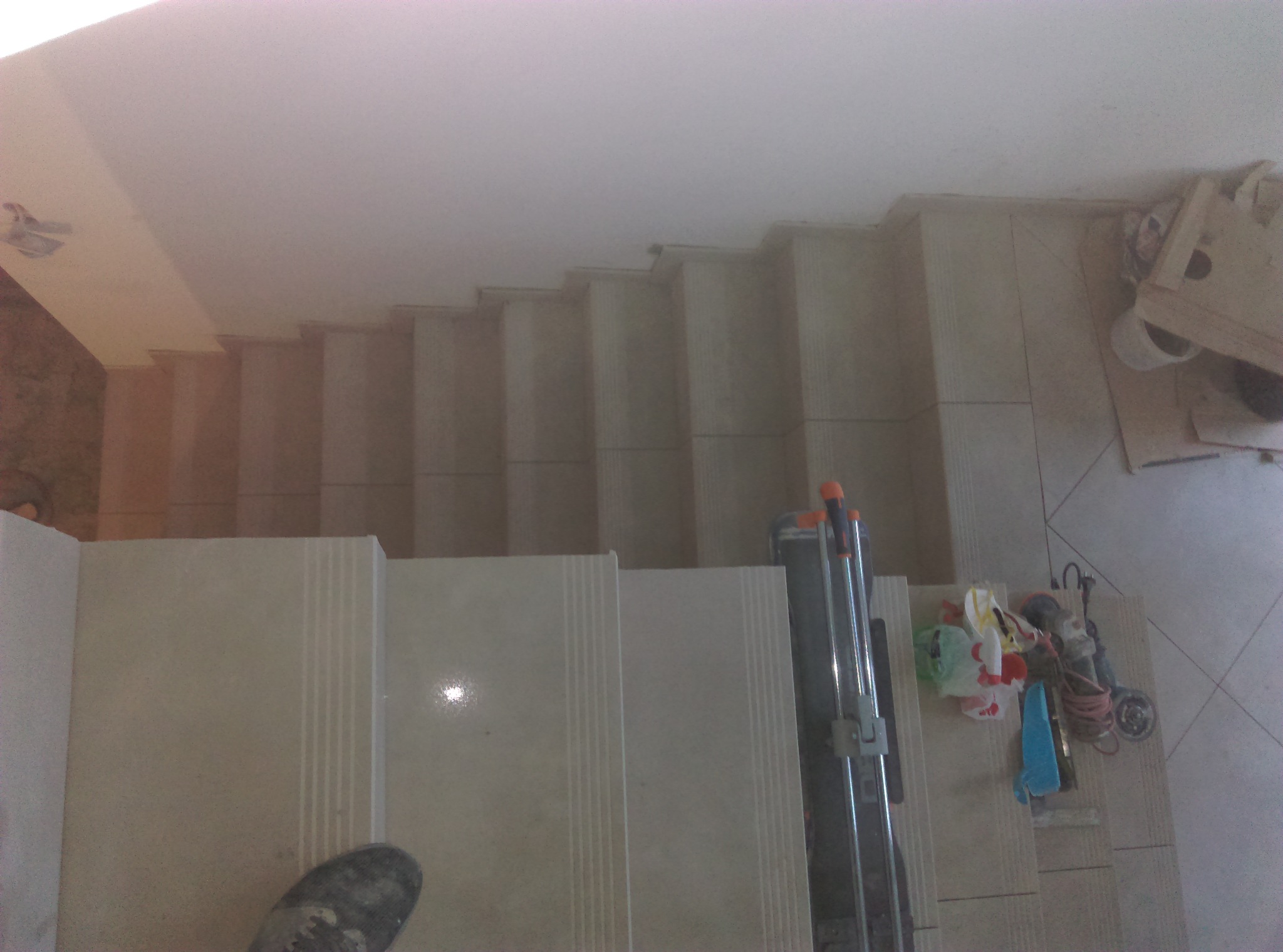 Schody wyłożone jasnymi płytkami, widok z góry, obok narzędzia glazurnicze i fragment buta