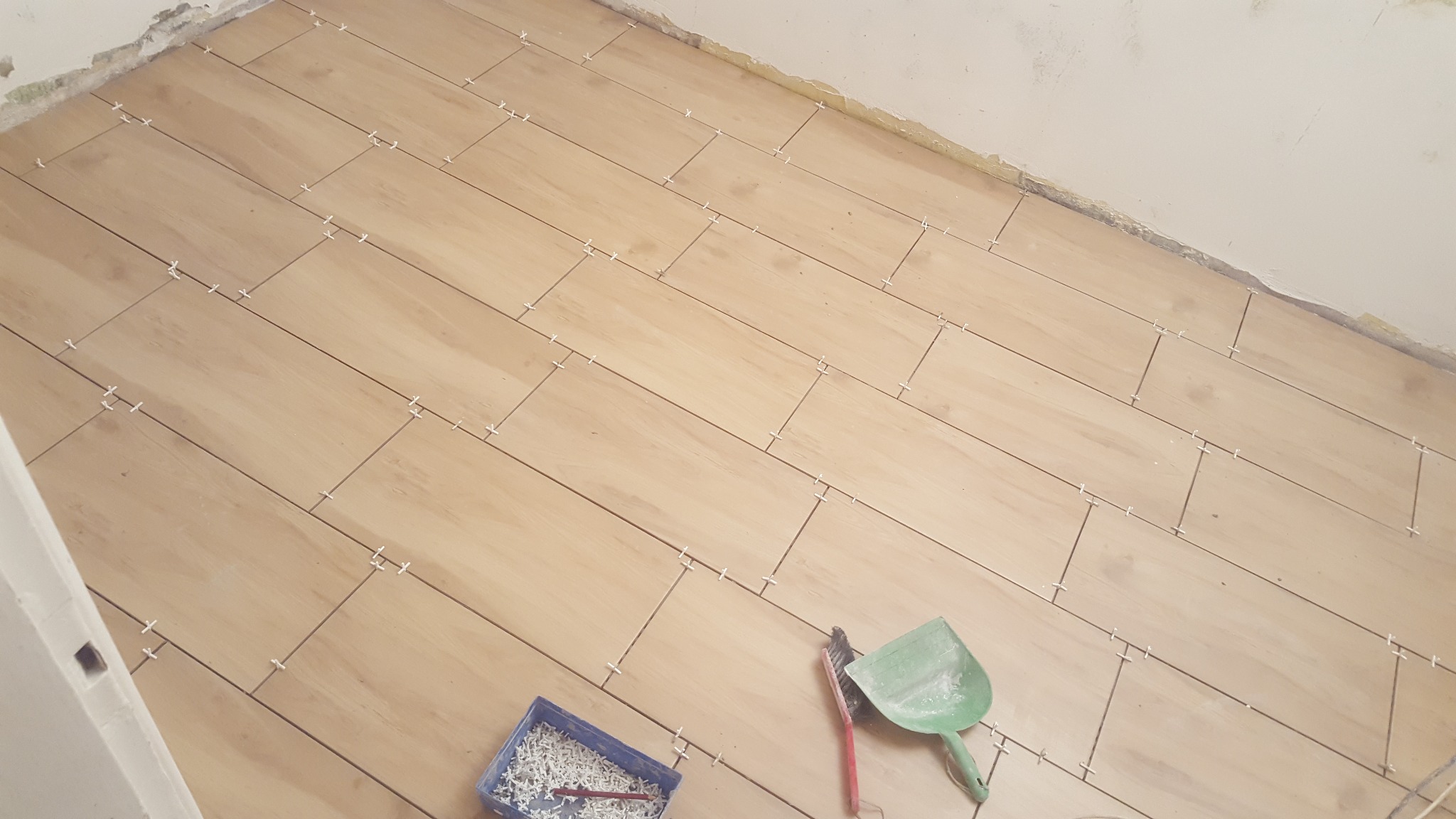 Ułożone płytki podłogowe imitujące drewno, z krzyżykami dystansowymi, widoczny fragment ściany w trakcie prac wykończeniowych, kuweta z krzyżykami i narzędzia do fugowania.