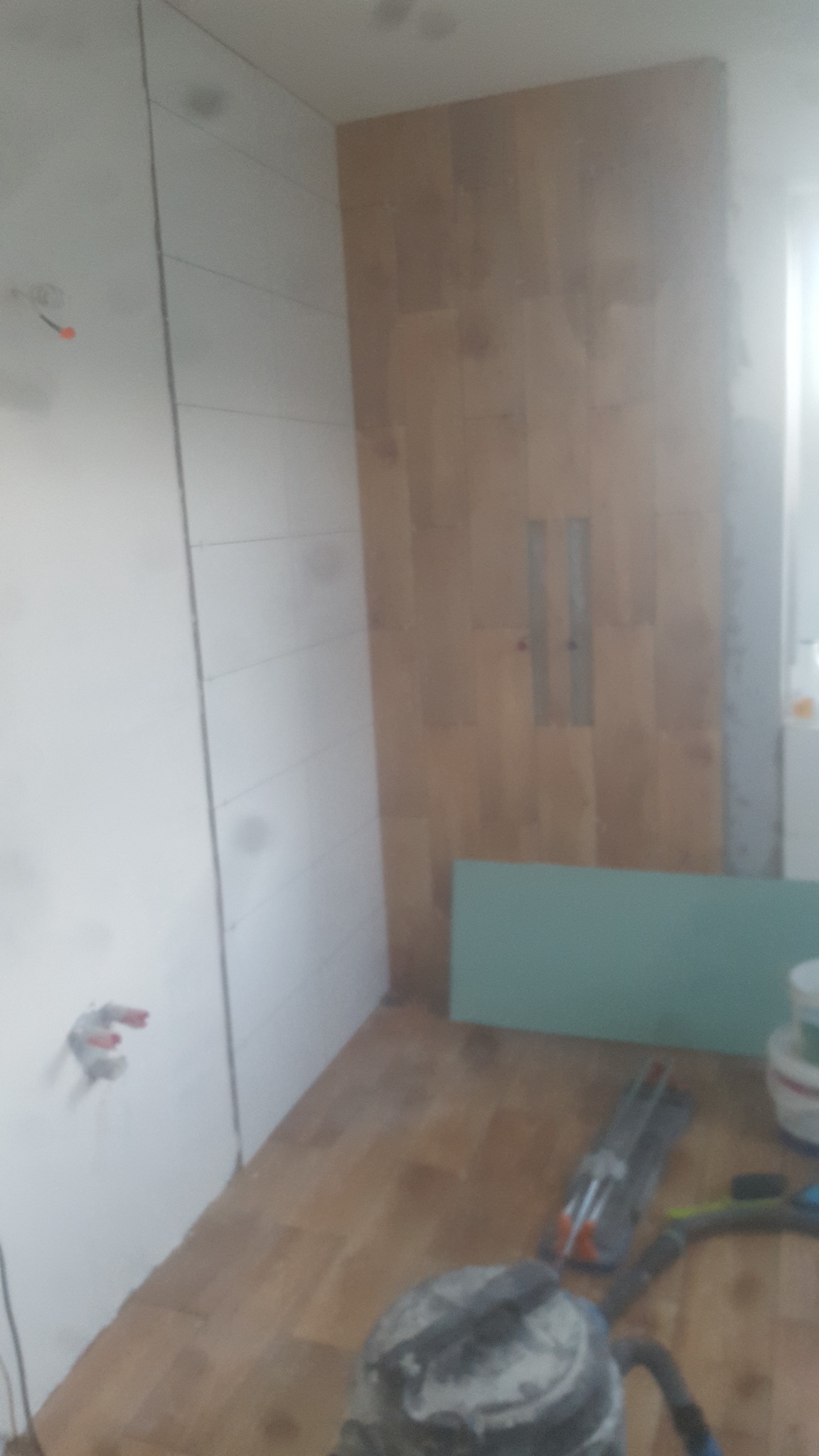 Fragment łazienki w trakcie prac wykończeniowych: częściowo ułożone białe płytki na ścianie, drewnopodobne płytki na drugiej ścianie z widoczną instalacją prysznicową, narzędzia glazurnicze...