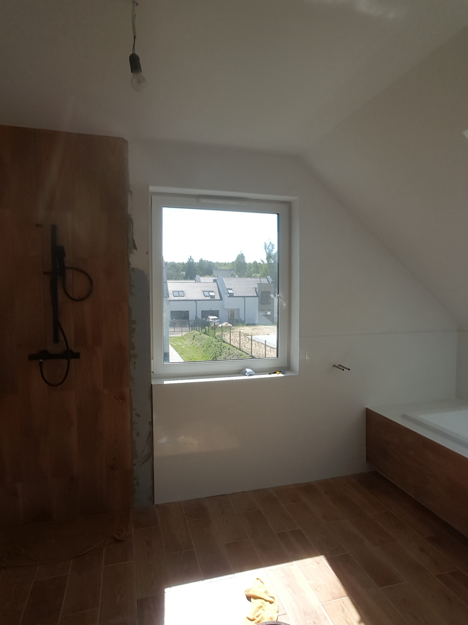 Wnętrze łazienki w trakcie wykończenia na poddaszu, widoczna drewnopodobna okładzina ściany z zamontowaną armaturą prysznicową, okno z widokiem na osiedle oraz drewniana zabudowa wanny, podłoga...