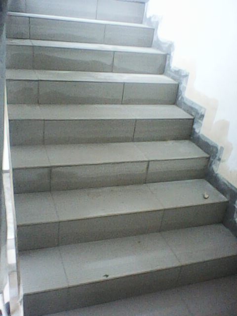 Wyłożone szarymi płytkami schody wewnątrz budynku, widoczne ślady po kleju na ścianie i stopniach.