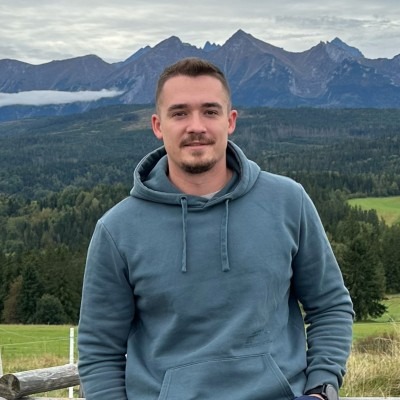 Mężczyzna w bluzie z kapturem na tle górskiego krajobrazu. Widoczne Tatry i lasy. Uśmiechnięty, z krótkim zarostem. Naturalne światło.