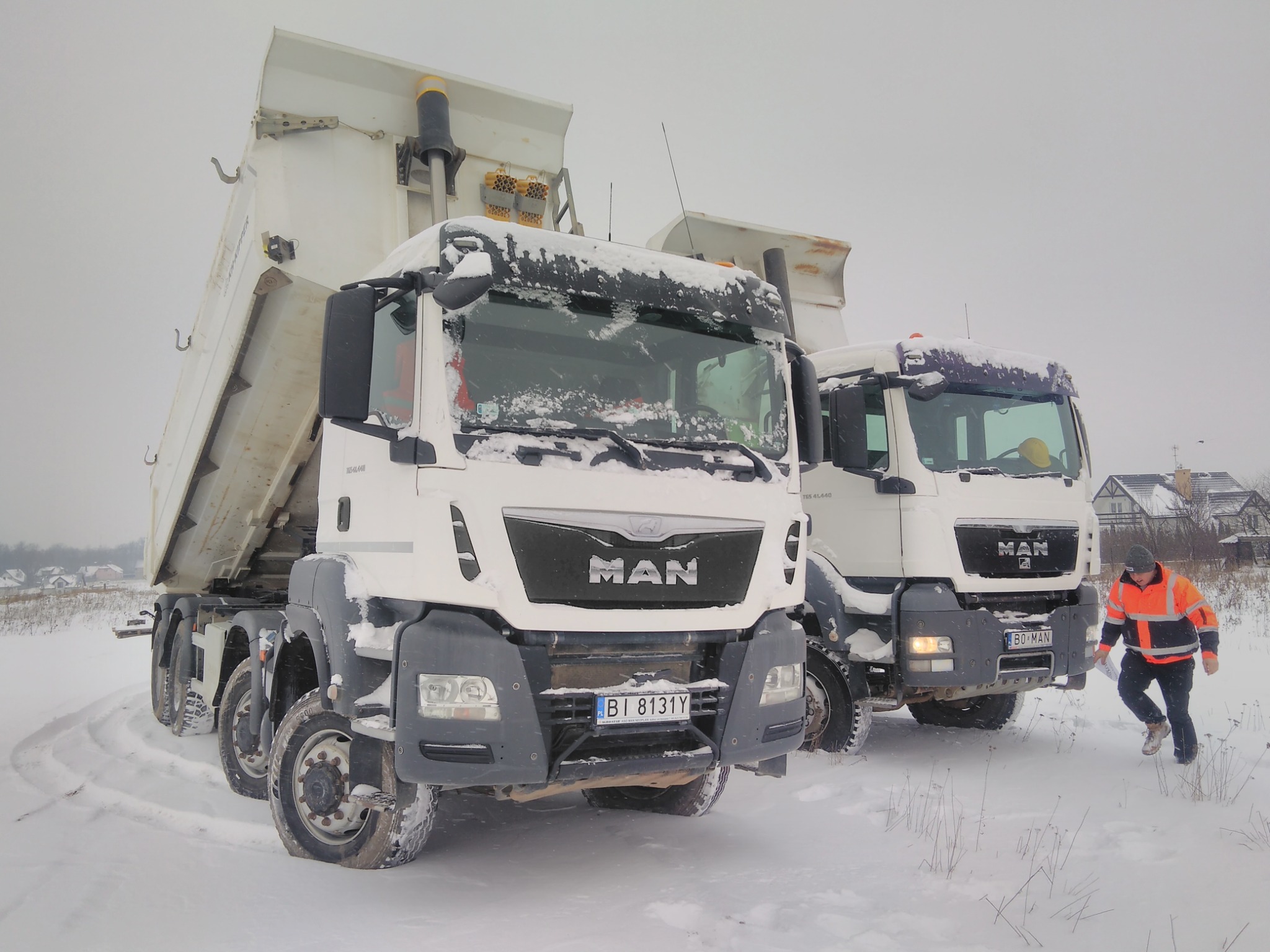 Dwa białe wywrotki MAN stoją na ośnieżonym terenie; jedna z podniesioną skrzynią ładunkową, pracownik w odblaskowej kurtce zbliża się do drugiego pojazdu.