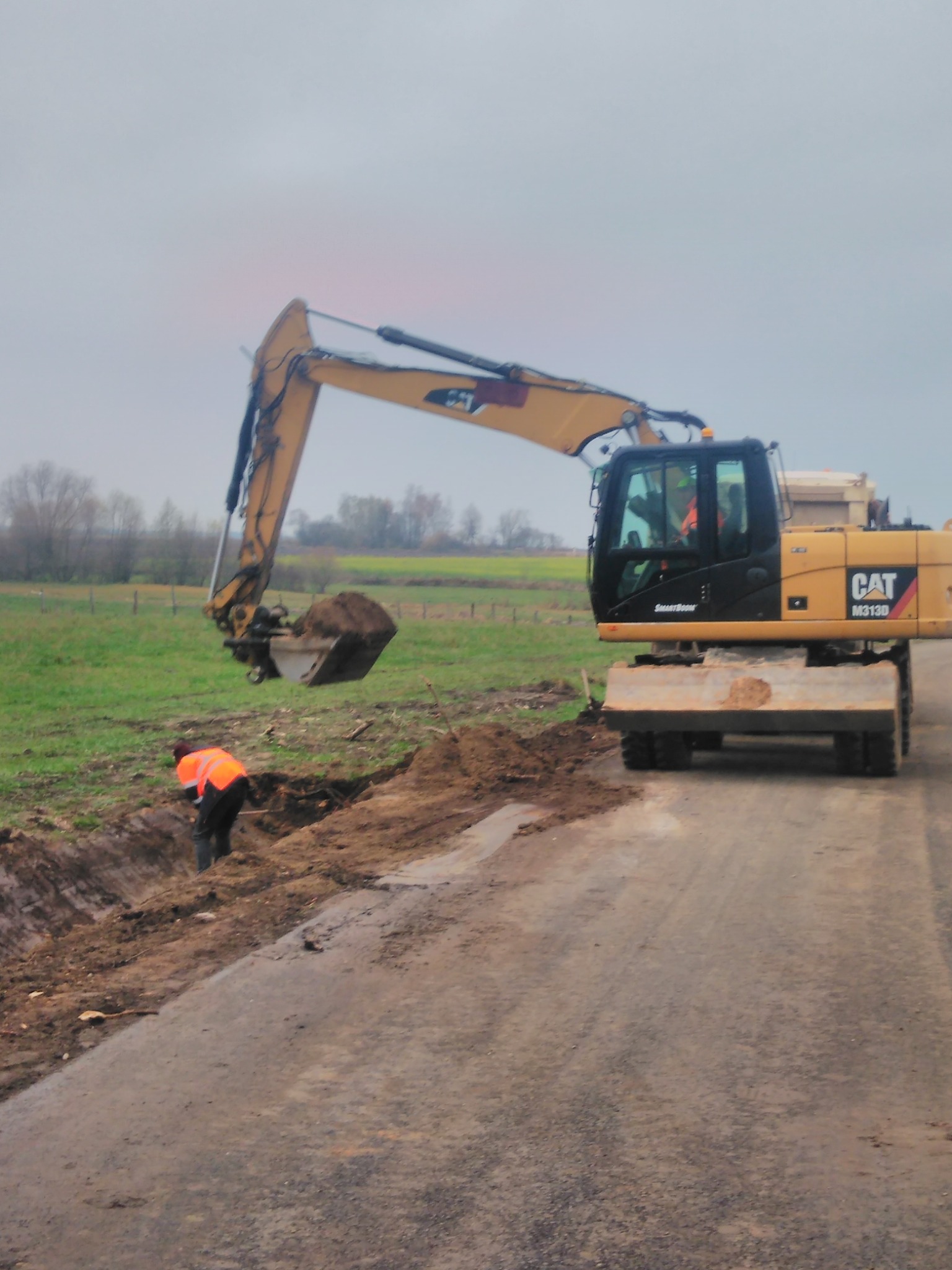 Żółta koparka CAT M313D z łyżką pełną ziemi, obok pracownik w pomarańczowej kamizelce przy wykopie wzdłuż asfaltowej drogi, w tle zielone pole i drzewa pod pochmurnym niebem.
