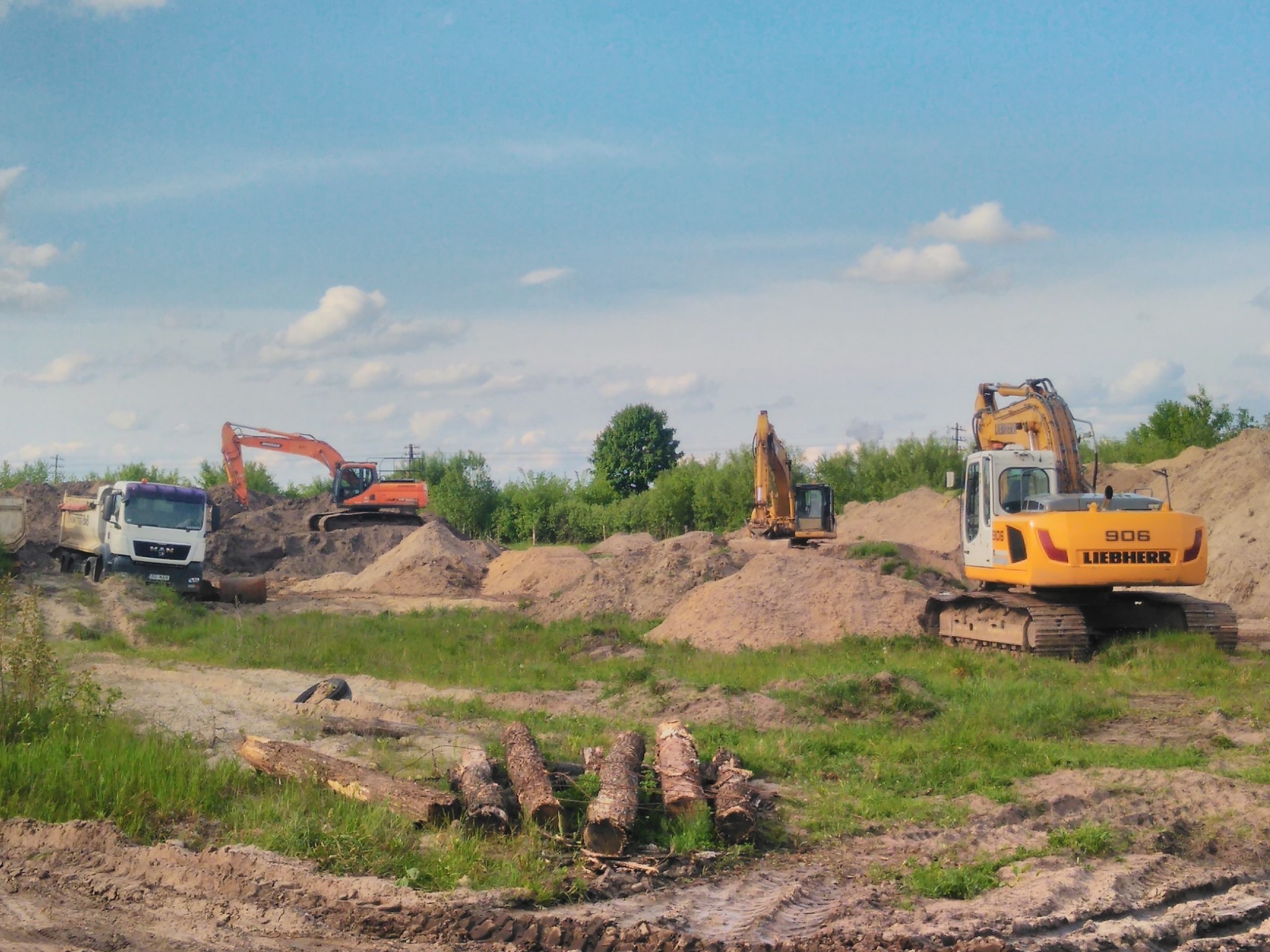 Żółty i pomarańczowy sprzęt budowlany (koparki i wywrotka MAN) na placu budowy z hałdami piasku i stosem kłód na pierwszym planie.