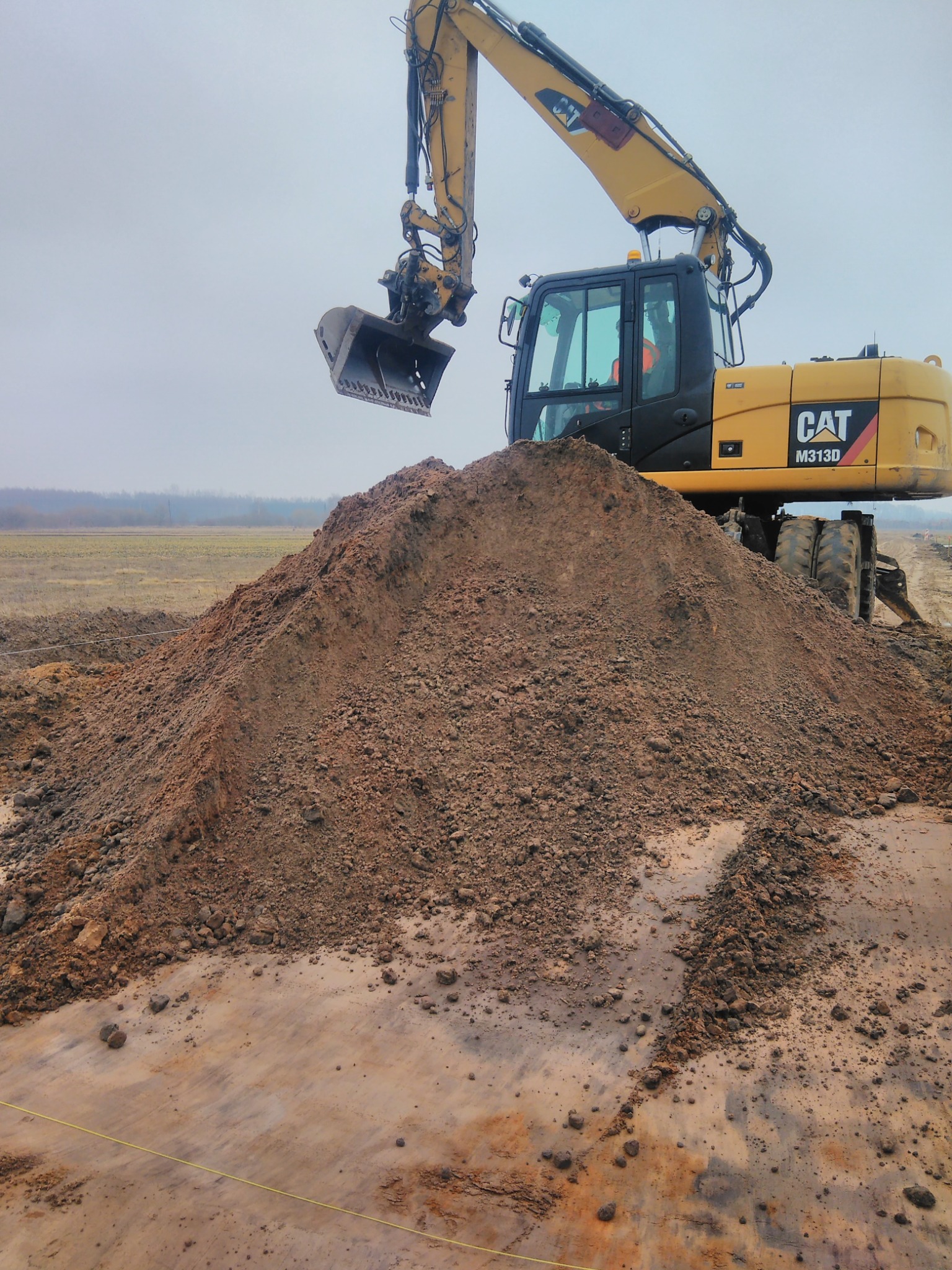 Żółty kołowy koparko-ładowarka CAT M313D z łyżką nad stertą ziemi na placu budowy w pochmurny dzień.