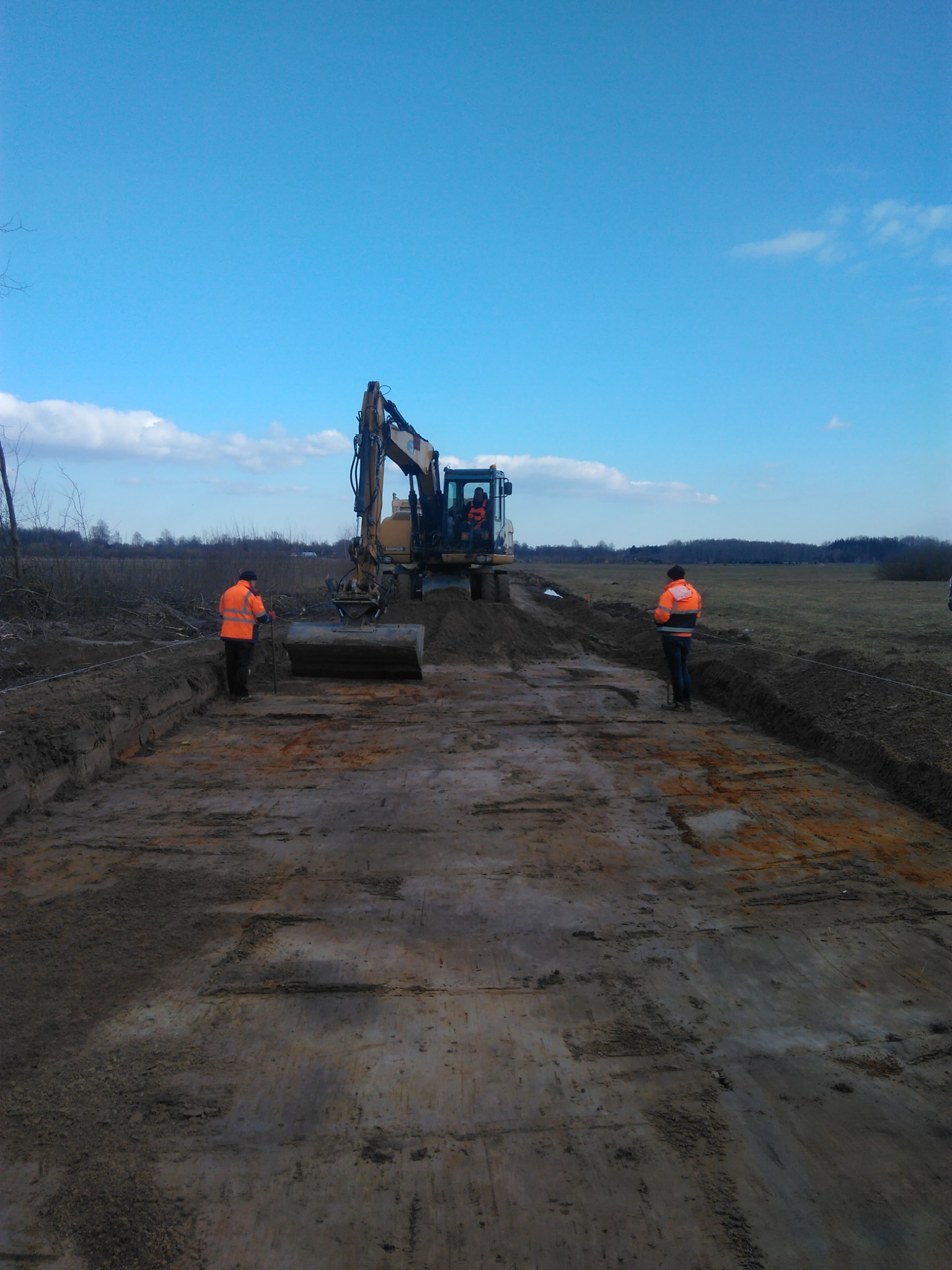 Żółta koparka na placu budowy, dwóch pracowników w pomarańczowych kamizelkach nadzoruje prace ziemne, wykop pod przyszłą drogę lub fundamenty.
