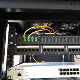 Panel krosowniczy światłowodowy z zielonymi złączami, zamontowany w szafie rack, nad przełącznikiem sieciowym Fortinet, z widocznymi wentylatorami chłodzącymi.