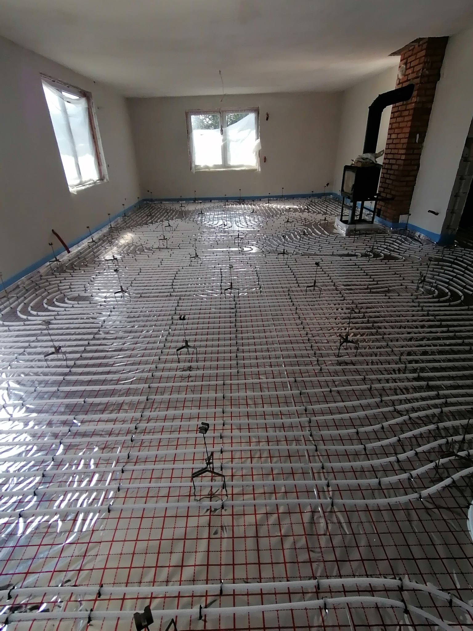 Rozłożone rury ogrzewania podłogowego na folii z siatką, widoczne uchwyty mocujące, w tle piec wolnostojący i surowe ściany w trakcie remontu.