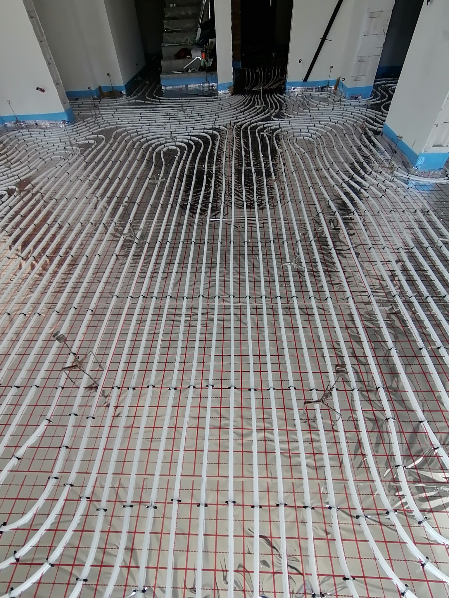 Białe rury ogrzewania podłogowego ułożone na folii izolacyjnej z czerwoną siatką, widoczne elementy montażowe i fragment schodów w tle.