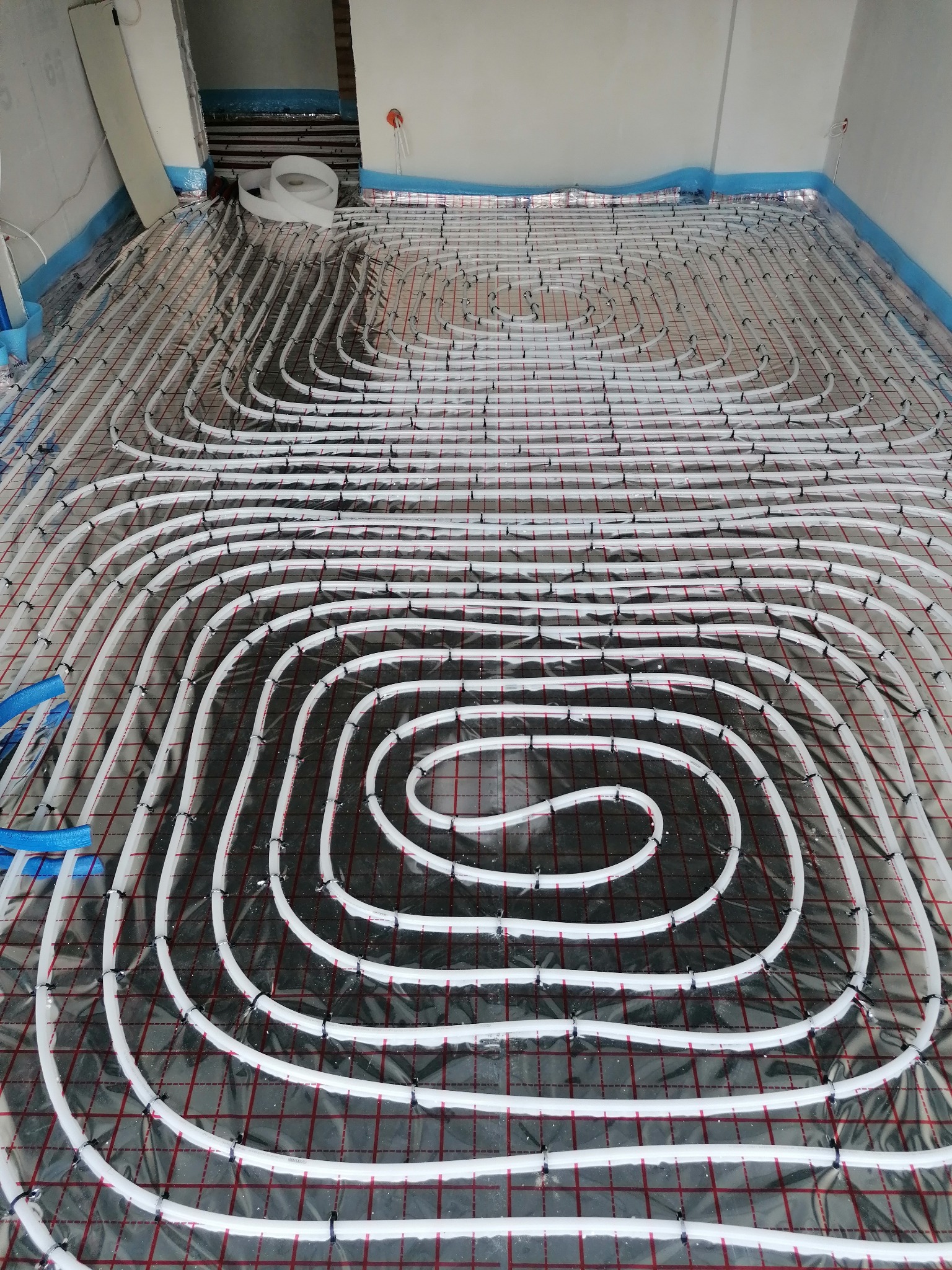 Białe rury ogrzewania podłogowego ułożone na folii z czerwoną siatką, przymocowane czarnymi klipsami w surowym wnętrzu budynku.