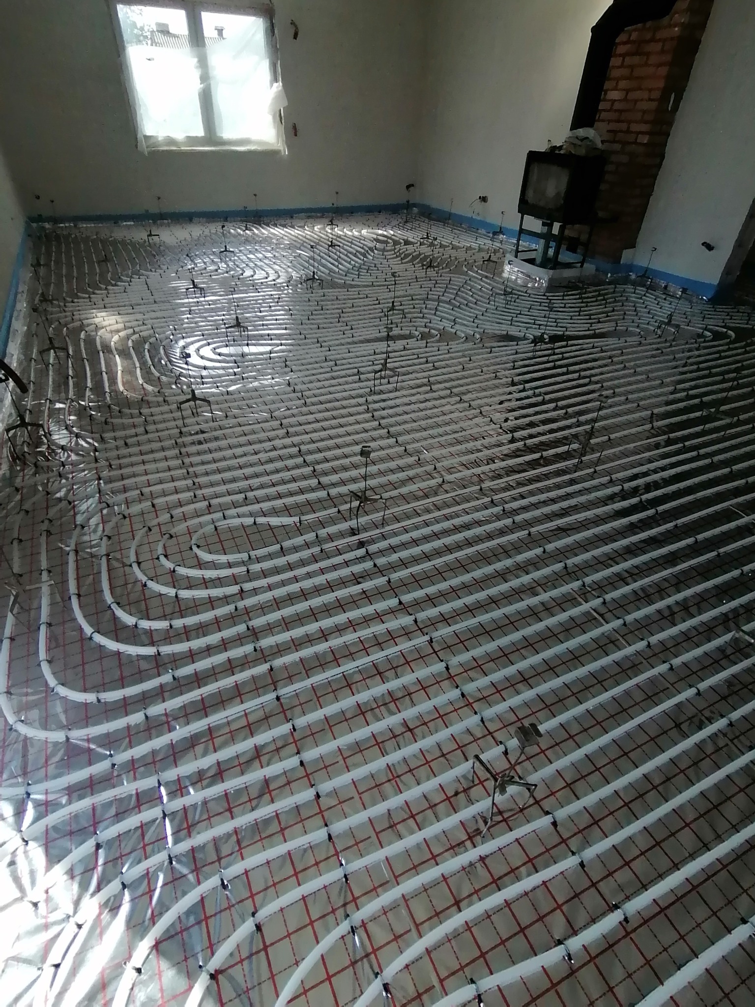 Białe rury ogrzewania podłogowego ułożone na folii z czerwoną siatką w surowym pomieszczeniu, widoczne okno i fragment kominka.