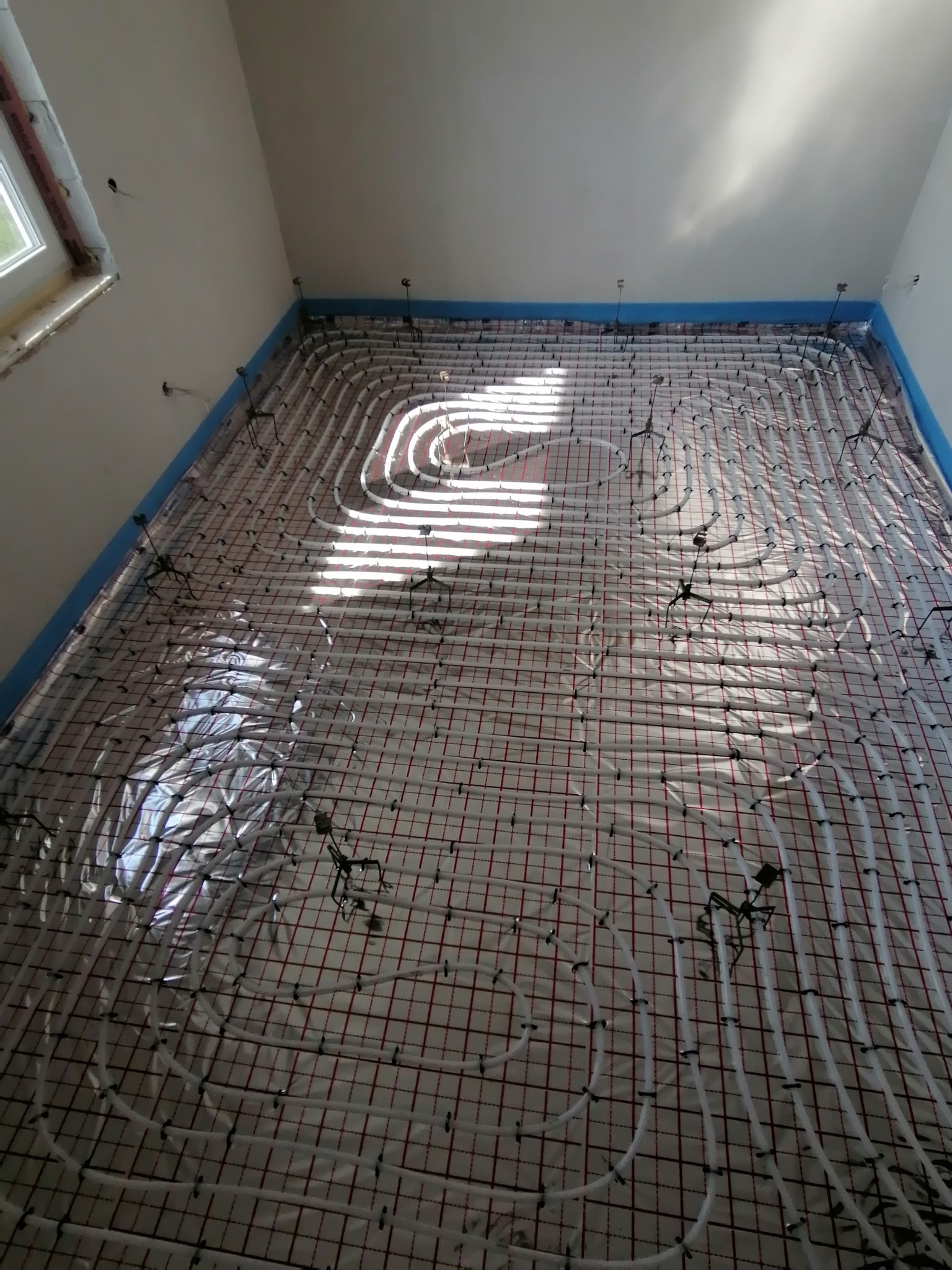 Białe rury ogrzewania podłogowego ułożone na folii termoizolacyjnej z czerwoną siatką, zamocowane czarnymi uchwytami, widok z góry w jasnym pomieszczeniu z oknem.
