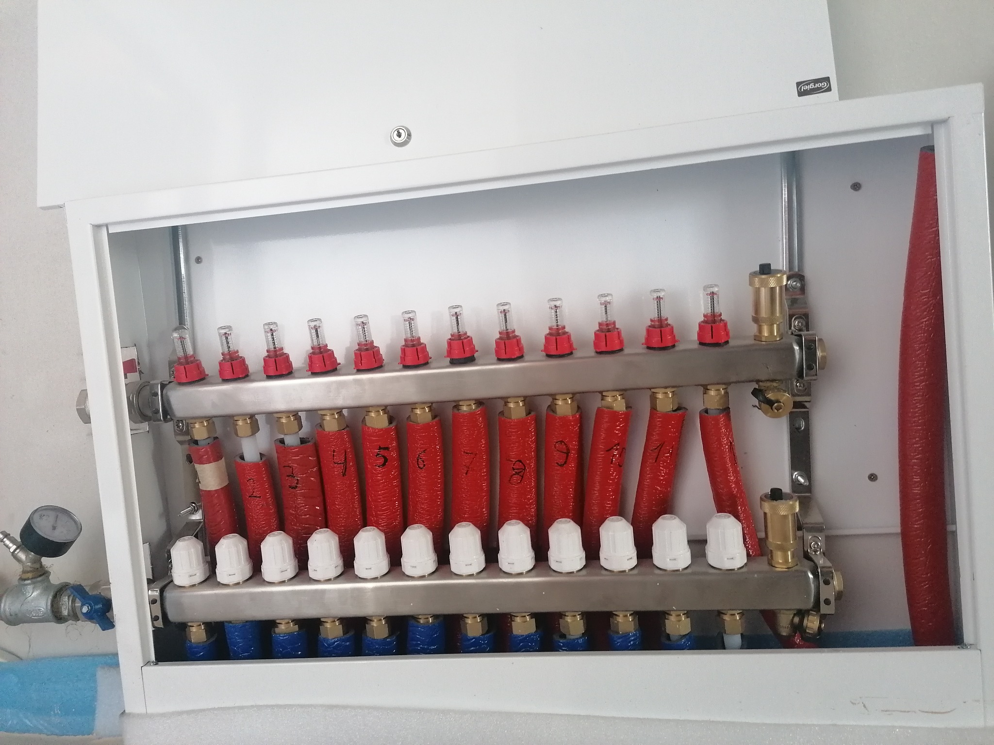 Rozdzielacz ogrzewania podłogowego w białej szafce, z widocznymi rurami grzewczymi oznaczonymi numerami od 4 do 10, manometrem i zaworami.