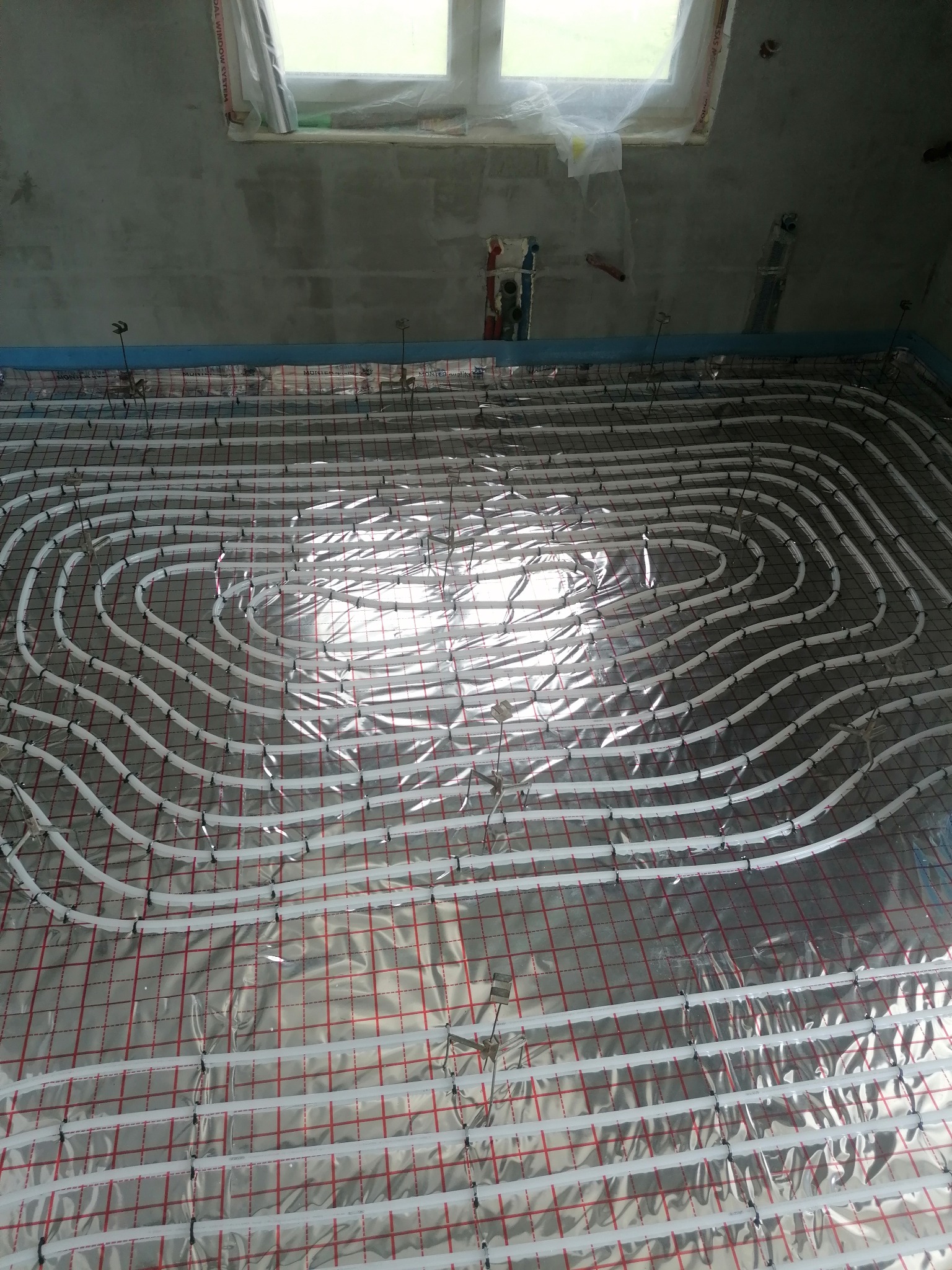 Białe rury ogrzewania podłogowego ułożone spiralnie na folii z czerwoną kratką, przymocowane do siatki metalowymi klamrami, widok z góry na surowy beton w tle.