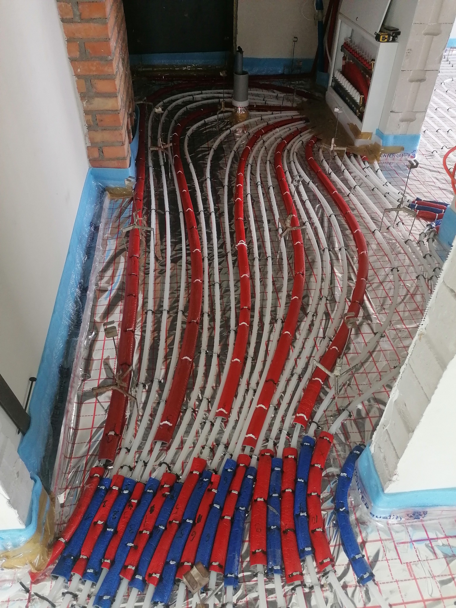 Rozłożone rury ogrzewania podłogowego na folii izolacyjnej z widoczną rozdzielnicą i fragmentem ceglanego łuku.