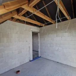 Surowy stan wnętrza poddasza z widoczną konstrukcją drewnianą dachu, ścianami z bloczków, wystającymi przewodami elektrycznymi i rurami instalacji wodnej w podłodze, przygotowane do dalszych prac...