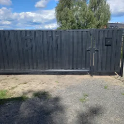 Przesuwna brama metalowa w kolorze grafitowym z inicjałami G i N, zamontowana między betonowymi słupkami, z widoczną skrzynką na listy i żwirowym podjazdem.