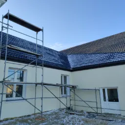 Nowy, ciemny dach z metalowej dachówki na jasnym budynku w trakcie budowy, widoczne rusztowania i lekki śnieg na dachu i ziemi.