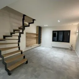 Nowoczesne schody wspornikowe z drewnianymi stopniami i czarną metalową konstrukcją w minimalistycznym wnętrzu z szarą podłogą, na ścianie półki z drewnianym fornirem.