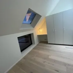 Jasne poddasze z drewnianą podłogą, oknem dachowym, wbudowanym oknem w ścianie, wnęką z oświetleniem i białymi szafami.