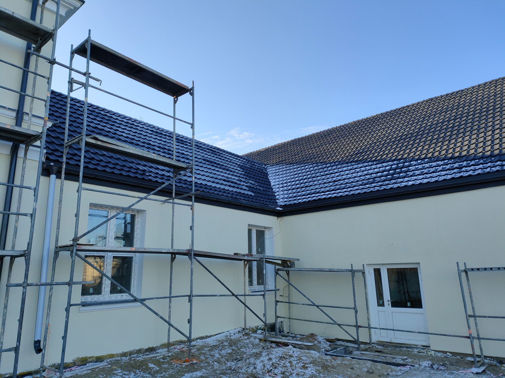 Nowy, ciemny dach z metalowej dachówki na jasnym budynku w trakcie budowy, widoczne rusztowania i lekki śnieg na dachu i ziemi.