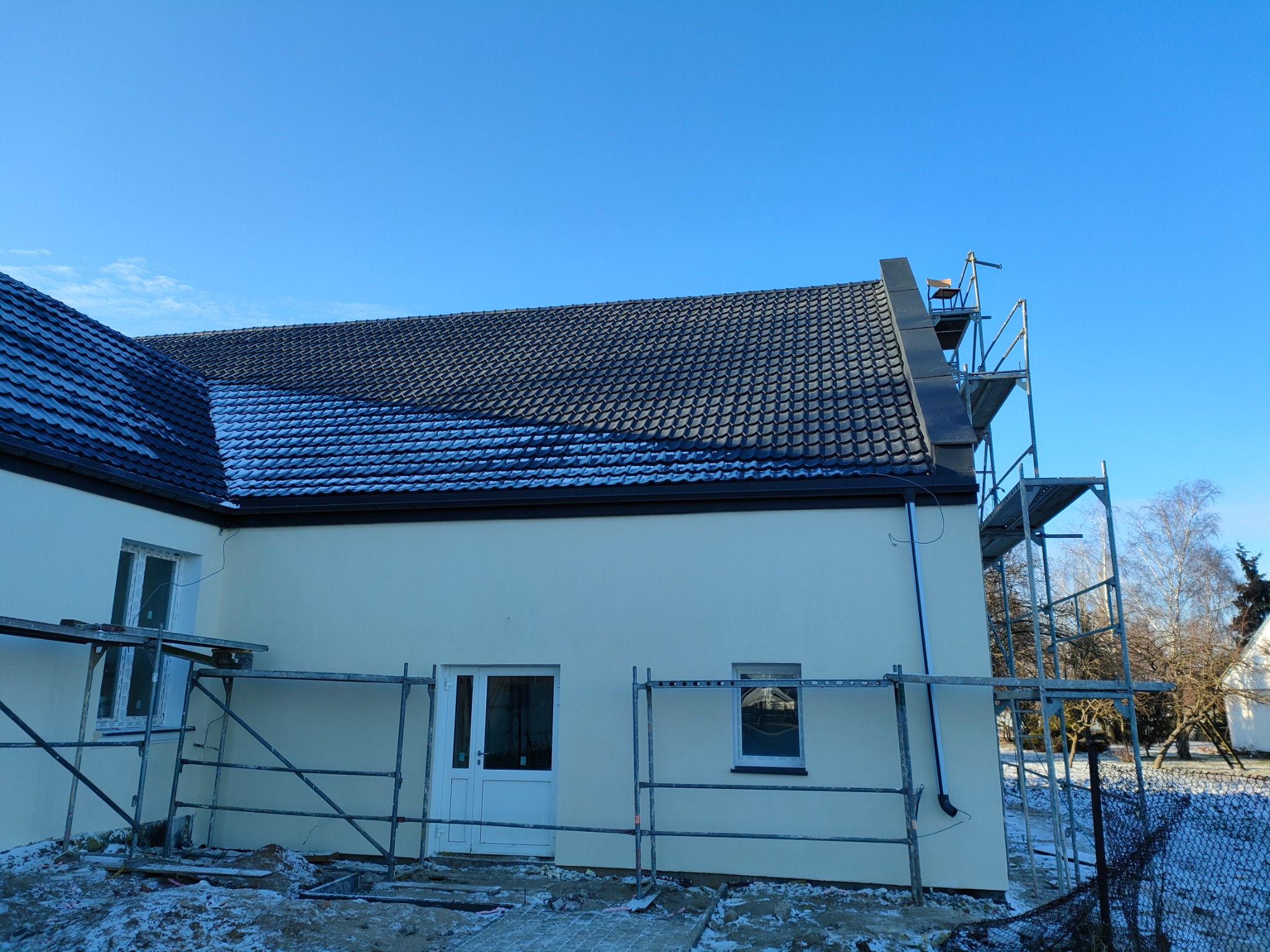Nowy dom jednorodzinny z ciemną dachówką, częściowo pokryty śniegiem, z rusztowaniem na dachu i przy ścianie, na tle błękitnego nieba w zimowej scenerii.