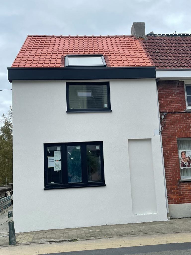 Dom z nową, czerwoną dachówką, oknem dachowym i czarną obróbką blacharską, przylegający do budynku z cegły.