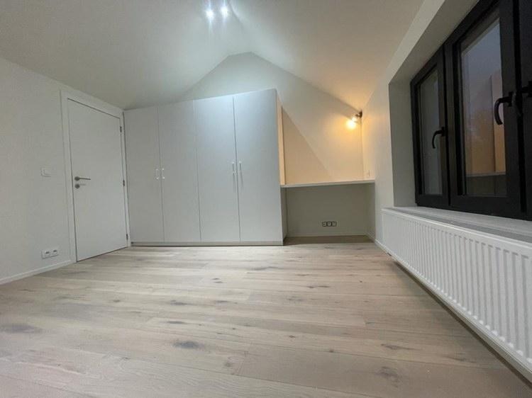 Poddasze po remoncie z jasną drewnianą podłogą, białymi ścianami, zabudową szafy i biurkiem pod skosem dachu, czarne okno z grzejnikiem pod spodem.