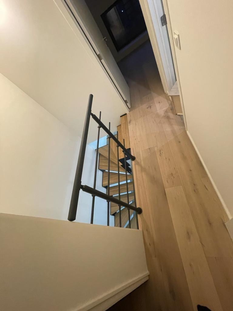 Spiralne schody z drewnianymi stopniami i czarną metalową balustradą, widziane z góry, z widokiem na drewnianą podłogę i okno dachowe w tle.