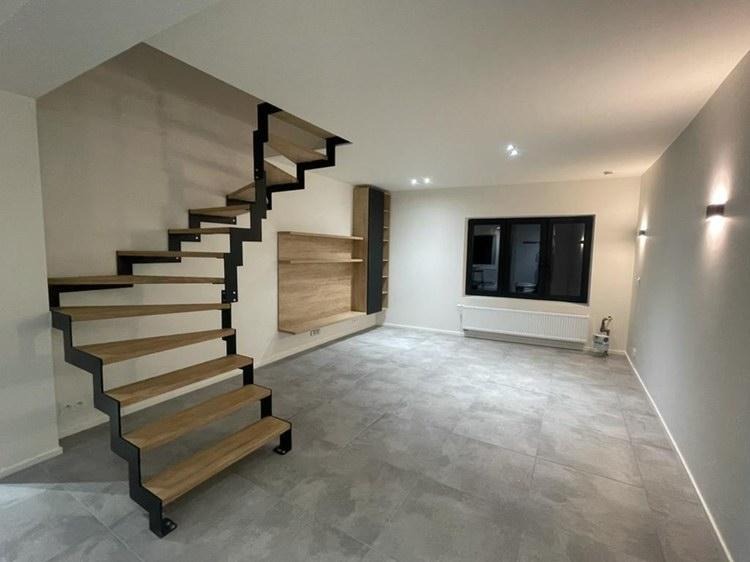 Nowoczesne schody wspornikowe z drewnianymi stopniami i czarną metalową konstrukcją w minimalistycznym wnętrzu z szarą podłogą, na ścianie półki z drewnianym fornirem.