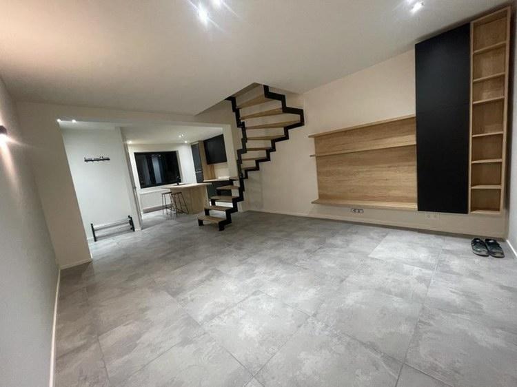 Wnętrze nowoczesnego domu z drewnianymi schodami spiralnymi z czarną metalową konstrukcją, jasnoszarą podłogą z płytek imitujących beton, zabudową ścienną z drewnianymi półkami i czarnym panelem...