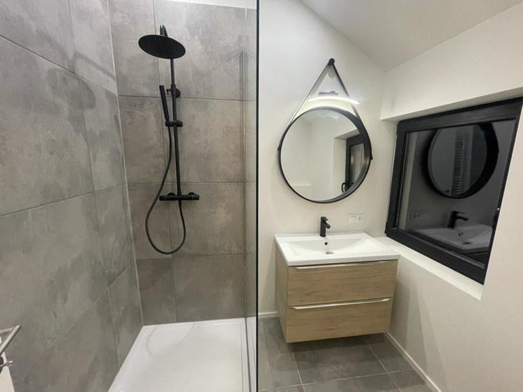 Nowoczesna łazienka z kabiną prysznicową wykończoną szarymi płytkami, czarną armaturą, okrągłym lustrem z podświetleniem LED zawieszonym na pasku i szafką pod umywalką z drewnopodobnym frontem.