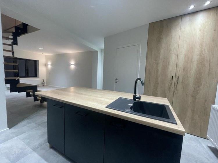 Nowoczesna kuchnia z wyspą: czarne szafki, blat imitujący drewno, czarny zlew i bateria. W tle schody ażurowe i minimalistyczne oświetlenie.