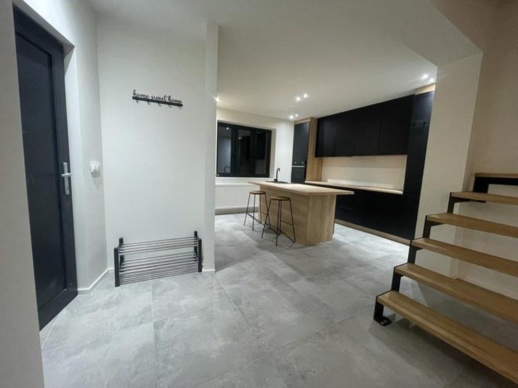 Nowoczesne wnętrze domu z szarymi płytkami podłogowymi, czarnymi meblami kuchennymi, drewnianym blatem i wyspą, metalowym regałem na buty przy wejściu oraz drewnianymi schodami z czarnymi elementami.