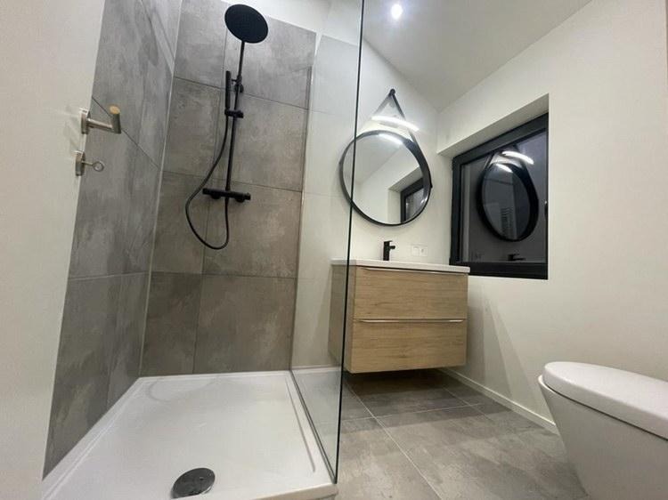 Nowoczesna łazienka z kabiną prysznicową typu walk-in, umywalką z szafką z jasnego drewna i okrągłym lustrem z podświetleniem LED w czarnej ramie.