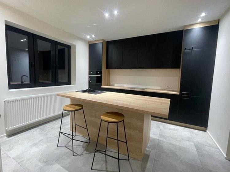 Nowoczesna kuchnia z czarnymi szafkami, drewnianym blatem i wyspą z dwoma hokerami na szarych płytkach podłogowych, oświetlona punktowymi lampami sufitowymi.