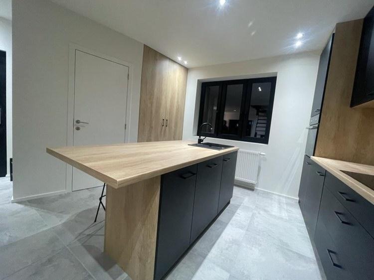 Nowoczesna wyspa kuchenna z blatem w kolorze jasnego drewna i czarnymi szafkami, czarna armatura, widok na okno i grzejnik, szara podłoga, minimalistyczny design.