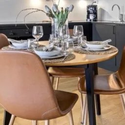 Stół z okrągłym blatem i czterema brązowymi krzesłami w nowoczesnej kuchni z czarnymi szafkami i drewnianą podłogą.