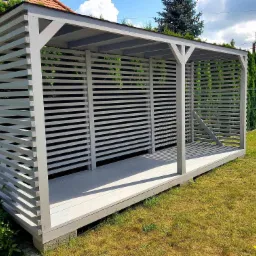 Szara, drewniana altana z poziomymi listwami, stojąca na trawniku w słoneczny dzień.