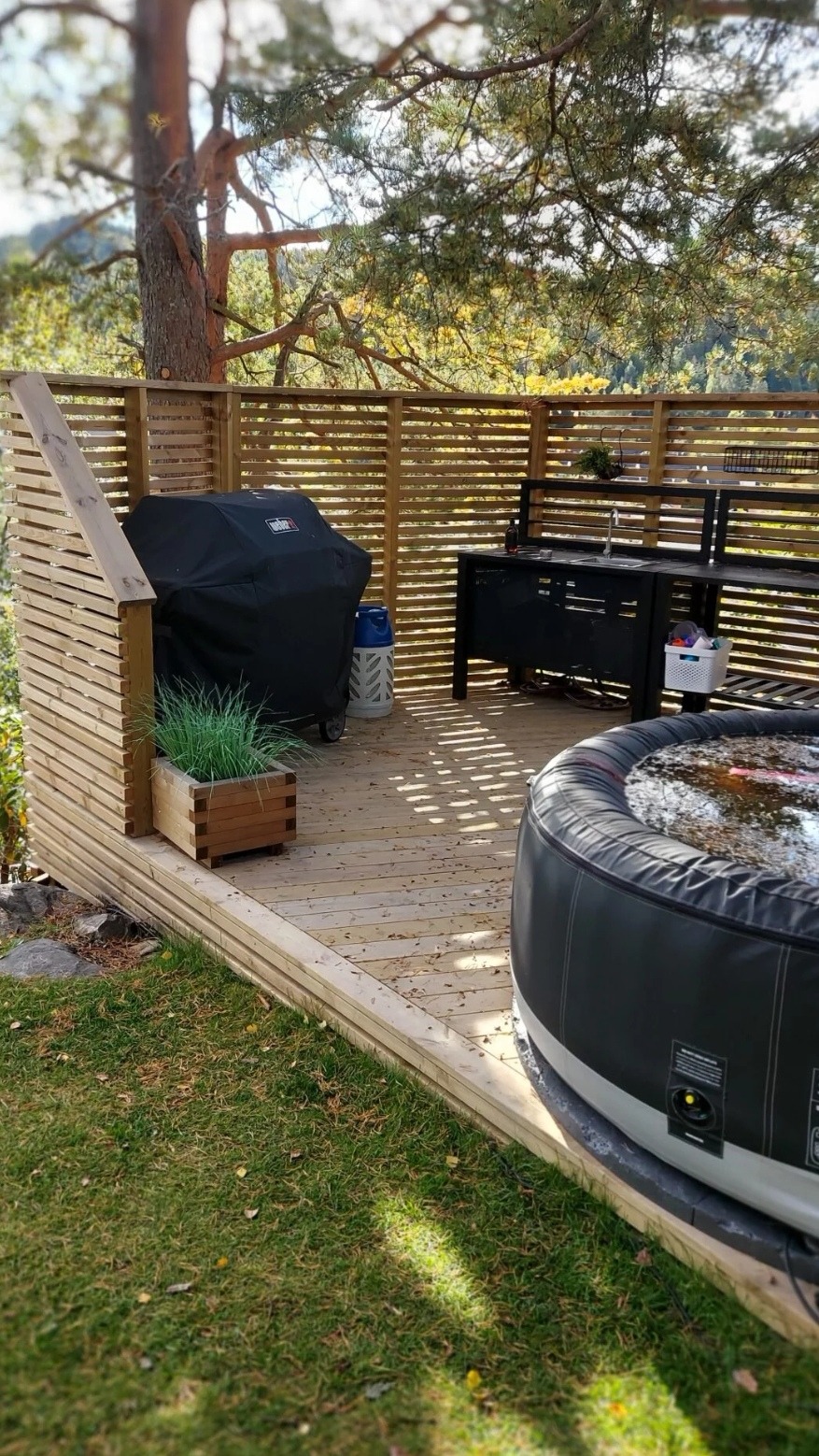 Drewniany taras z jacuzzi, grillem pod pokrowcem i zabudową z poziomych desek, otoczony zielenią.