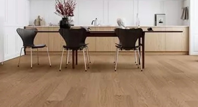 Jasna, drewniana podłoga w jadalni z trzema czarnymi krzesłami o metalowych nogach ustawionymi wokół ciemnego stołu; w tle białe ściany i jasne szafki kuchenne z dekoracjami.