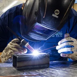 Spawanie metodą TIG rury kwadratowej przez spawacza w masce z logo Miller, iskry i jasne światło w miejscu spawania.