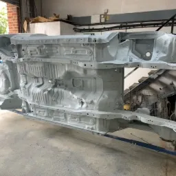 Podwozie samochodu po oczyszczeniu i pomalowaniu farbą podkładową, umieszczone na wózku warsztatowym, widok od spodu, w tle brama garażowa i schody.